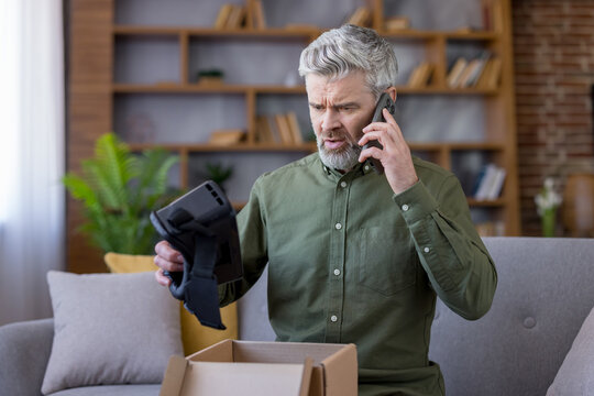 Mature man holding a new virtual reality headset with a perplexed expression, making a phone call, expressing dissatisfaction with unboxing a defective product at home - Powered by Adobe
