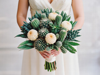 Close-up of the bride's hands with a bouquet. Bridal bouquet combines spiny barrel cacti, rosette succulents, creamy preserved blooms, white ribbon, contrasted textures against wedding gown