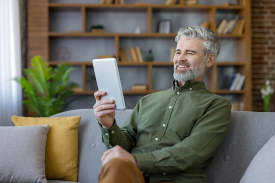 Mature man sitting comfortably on a sofa at home, enjoying leisure time while using a digital tablet for communication or entertainment, embodying modern senior lifestyle
