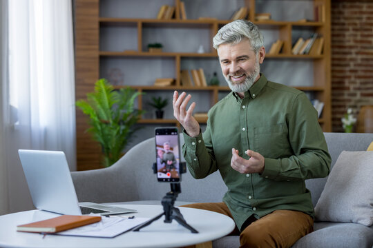 Senior man sitting on a sofa in his home office actively recording a video for a vlog or webinar using a smartphone, on a tripod while looking at the camera and gesturing - Powered by Adobe