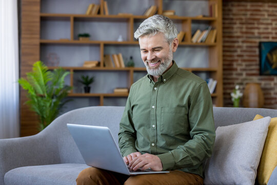 Smiling senior man with gray hair and beard sitting on a comfortable sofa, confidently engaging in remote work using a laptop in his modern living room