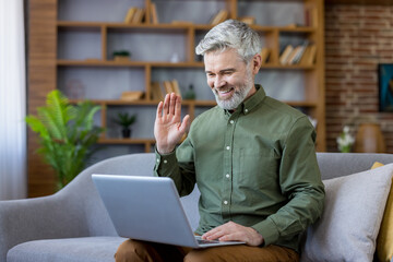 Smiling mature man with grey hair and beard having a video conference on his laptop, sitting comfortably on a couch at home, engaging in virtual communication and connection