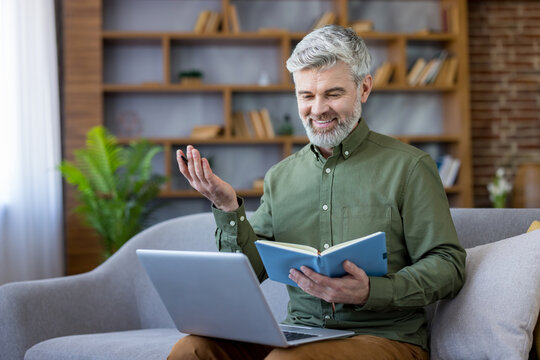 Mature man with a silver beard conducting an online video conference while sitting on a sofa at home, holding a notebook and gesturing during conversation