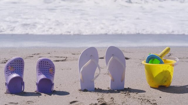 beach flip-flops and child&rsquo;s purple clogs standing in sand with ocean waves in background, family beach vacation, and summer joys, sandals rest side-by-side on shore, parenting, childhood memories