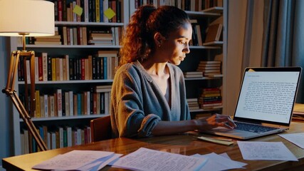 woman at desk working late with laptop and scattered papers under warm lamp light, researcher typing notes,