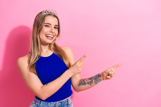 Smiling young woman with blonde hair wearing a blue top and tiara stands against a pink background pointing to the side for fashion lifestyle and shopping promotion