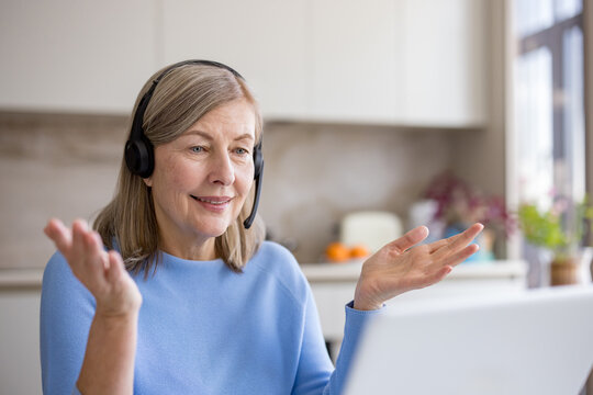Senior woman actively participating in a virtual meeting from home, communicating through a headset with microphone while working remotely on a laptop, embracing modern technology for connection