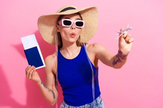 Young woman in a blue tank top and straw hat promotes travel and shopping with a toy airplane against a bright pink background - Powered by Adobe