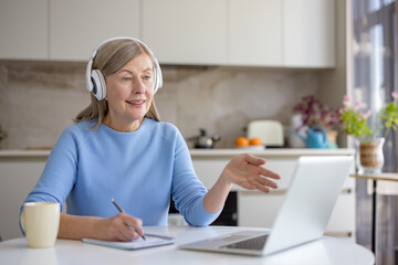 Senior woman with headphones on a kitchen table, engaged in an online video call gesturing, taking notes on a laptop while discussing and learning remotely at home