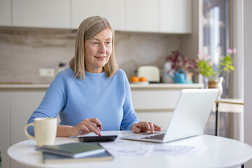 Mature woman at kitchen table managing household finances balancing bills, calculator and laptop, focused on budgeting, retirement planning and online accounting in a comfortable home setting
