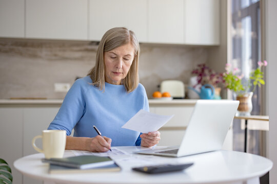 Mature woman reviewing financial documents and making notes while using a laptop and calculator for budget planning and remote work from her modern kitchen