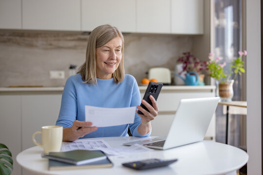 Senior woman is managing her personal finances and paying bills online using a smartphone and laptop at home, smiling while organizing household expenses and financial records