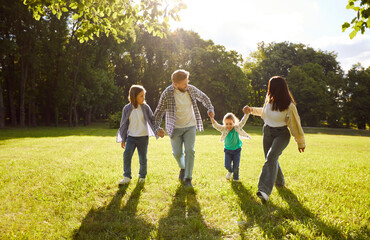 Fototapeta premium Four family members having fun at park during sunny summer day, walking together and holding hands, having good relationships, enjoying nice day. Parents and children resting outdoors