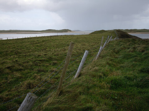rustic fence lines a grassy coastal field in County Donegal Ireland, leading to distant dunes under cloudy sky and strong side wind
