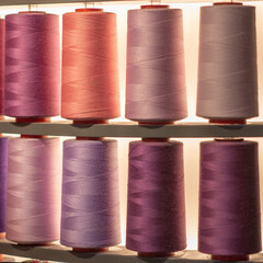 Close-up of numerous large thread spools arranged in a warm color gradient of reds, pinks, and creams. Organized textile manufacturing or sewing supply display with backlighting.