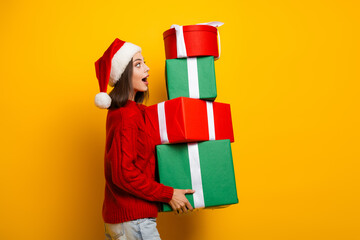 Funny holiday moment as pretty girl carries a tall stack of Christmas gifts on a bright yellow background during festive season