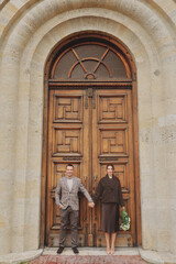Elegant couple holding hands and standing in front of a massive, ornate wooden door of an old stone church or historic building