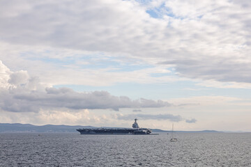 Fototapeta premium Aircraft Carrier On Calm Sea Under Cloudy Sky With Small Sailboat Nearby