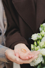 Close-up of a couple's hands holding two golden wedding rings over a bouquet of white roses, symbolizing proposal or marriage