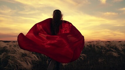 Little girl in red cape plays superhero on grass in park, sunset. Happy child runs across field in red superhero cape. Child winner plays in superhero costume in nature park. Game concept for children