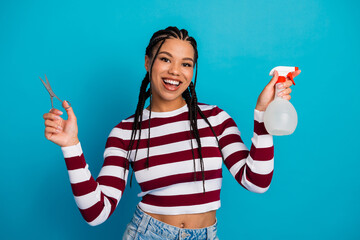 Young woman with striped top smiling holds scissors and spray bottle against blue background