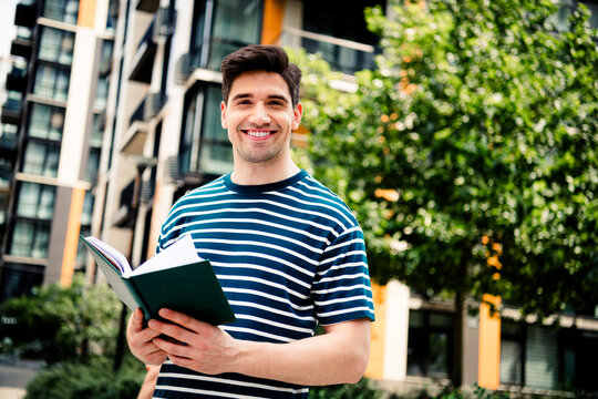 Smiling young man outdoors holding a book dressed casually in urban surroundings with modern architecture and greenery