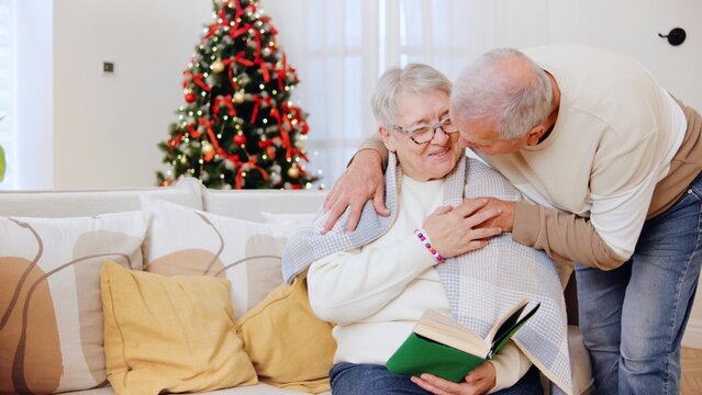An elderly couple enjoys a tender moment while reading together by a decorated Christmas tree, husband covers his wife with a blanket