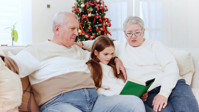 Grandparents reading a story to their granddaughter during a cozy New Year's celebration at home by the Christmas tree