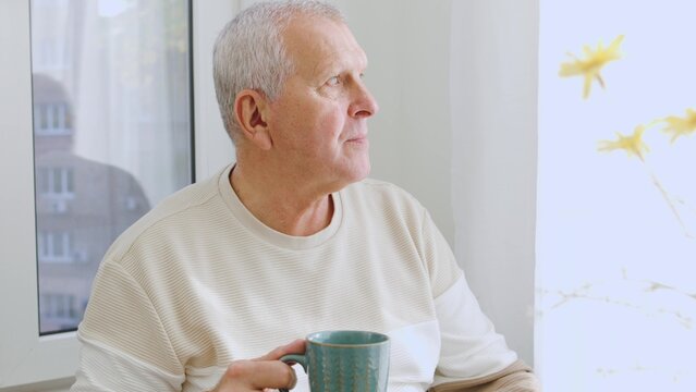 Grandfather is sitting by the window in his cozy house and drinking hot tea.