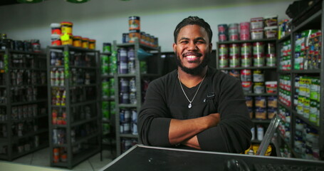 Man standing confidently at paint store counter with arms crossed, surrounded by paint cans and supplies, welcoming worker in professional retail setting