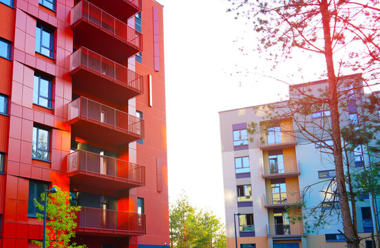 Colorful modern apartment buildings in sunny neighborhood with green trees in foreground. Sunlight shines on balconies and trees, creating cheerful atmosphere. Red facade of modern condominium.