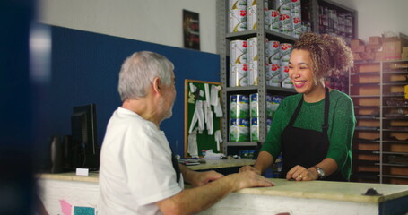 Young woman behind a counter smiling warmly while interacting with an older man in a hardware store...