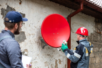 Two workers in overalls are setting up a satellite dish mounted on the wall of an old village house