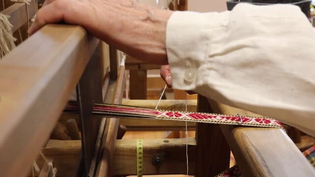 woman dressed in traditional Latvian attire is working on a wooden loom in a cozy room filled with weaving equipment. The scene captures the beauty of craftsmanship and cultural heritage, with colorfu