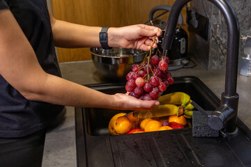 Person Washing Fresh Fruit Under Kitchen Faucet. Individual rinsing red grapes under running water with bananas, oranges, and plums soaking in the kitchen sink below.