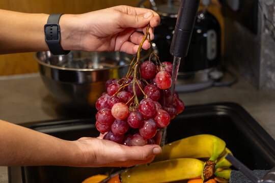 Washing Fresh Red Grapes in Kitchen Sink. Close-up of hands rinsing red grapes under running water in a kitchen sink, promoting healthy eating and food hygiene.