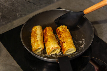 Cooking Cheese Blintzes in Frying Pan on Stove. Three golden cheese-filled blintzes frying in a pan with butter on a stove top, being flipped with a black spatula.