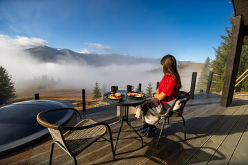 Fototapeta premium Woman Enjoying Scenic Breakfast on Mountain Deck. Woman having breakfast on a wooden terrace overlooking a foggy mountain landscape, wrapped in a blanket on a peaceful morning.