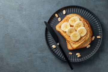 Peanut butter toast and banana on a black plate on a dark background with nuts and knife.