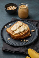 Peanut butter toast and banana on a black plate on a dark background with nuts