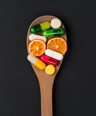 Wooden spoon filled with colorful vitamin capsules and dried citrus fruit slices on a black background representing the balance between natural food and supplements.