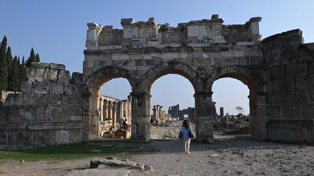 A woman walking through the Domitian Gate in the ancient city of Hierapolis. The view of the historical ruins and the bright sky under the beautiful sunset light.