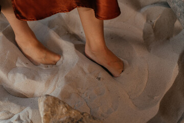 Woman in a satin skirt sitting barefoot on sand with natural dried plants and stones in a rustic boho setting