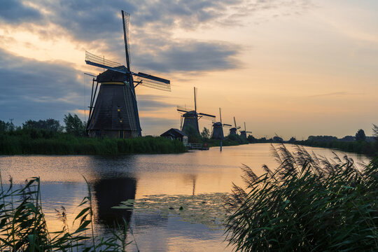 Traditionelle Windm&uuml;hlen stehen an einem ruhigen Kanal im Abendlicht. Der Himmel leuchtet in sanften Farben, die sich im Wasser spiegeln &ndash; eine stimmungsvolle Landschaft.