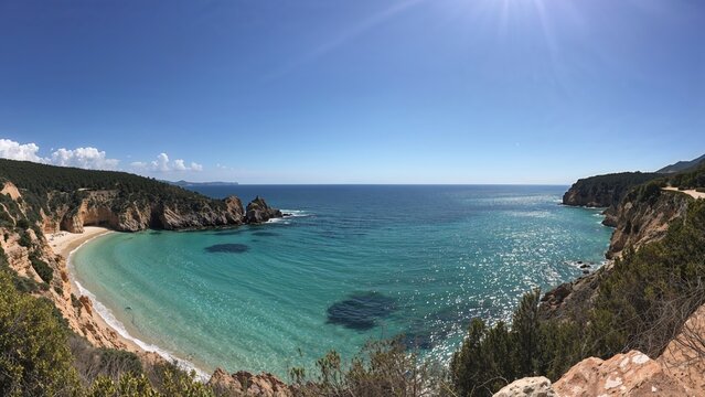 Coastal bay view from cliffside with blue sky and sun rays, featuring sandy beach and rocky cliffs. Nature scenery and ocean landscape.