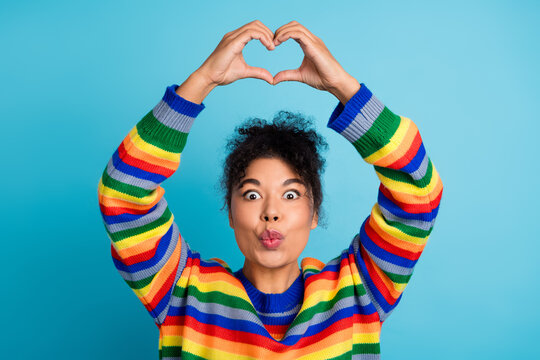 Young woman in rainbow striped sweater makes heart gesture against blue background smiling and playful lifestyle portrait