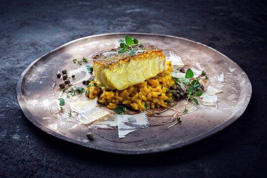 Fried nordic skrei cod white sea fish filet with safran risotto rice, green vegetable and parmesan cheese served as close-up on a rustic design plate