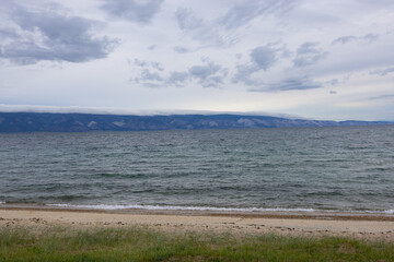 A serene view of Lake Baikal with calm waters reflecting the sky. Mountains and an island are visible in the distance under a cloudy sky.