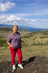 A young white woman sits on a rock near Lake Baikal. She is wearing a plaid shirt and red trousers.