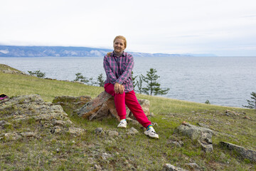 A young white woman sits on a rock near Lake Baikal. She is wearing a plaid shirt and red trousers.